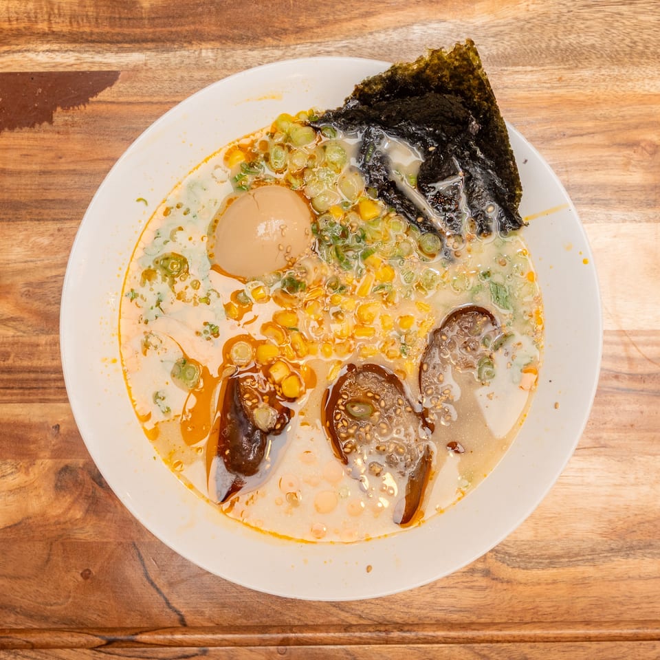 Top-down view of ramen in a white bowl, topped with egg, corn, scallions, seaweed, mushrooms, and chili oil.
