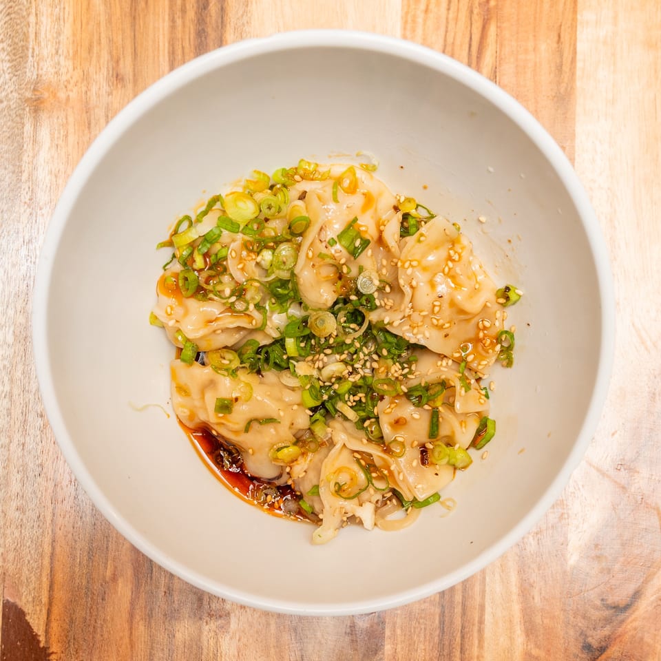 Bowl of dumplings topped with sliced green onions and sesame seeds in spicy chili oil on a wooden table.