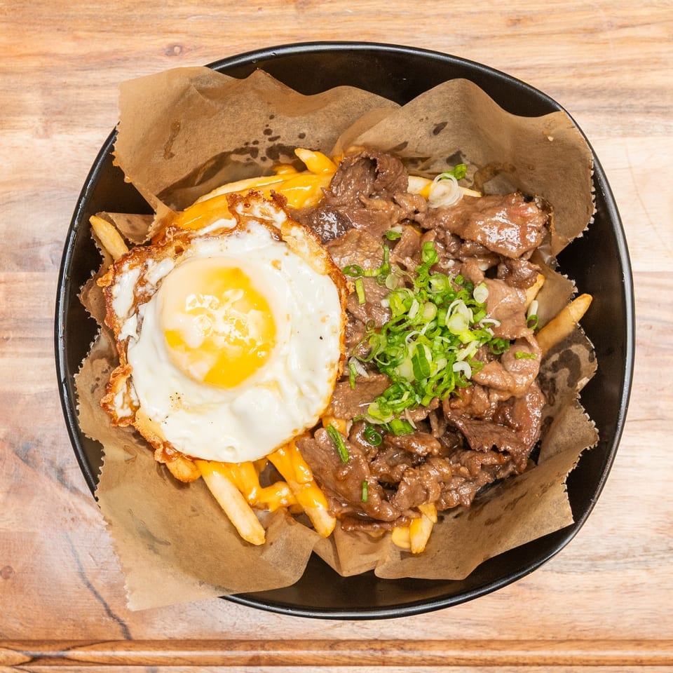 Loaded fries topped with melted cheese, sliced beef, a fried egg, and green onions in a black bowl.