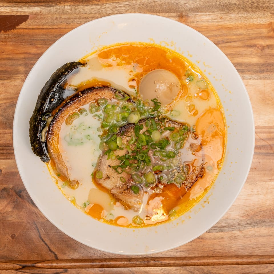 Top-down view of ramen in a white bowl with orange broth, sliced pork, egg, seaweed, and chopped green onions.