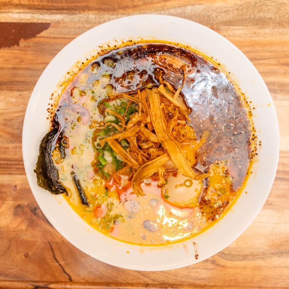 Top-down view of spicy noodle soup topped with crispy vegetable strips, scallions, chili oil, and seaweed on a wooden table.