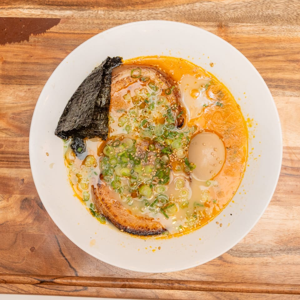 Overhead view of ramen in a white bowl with pork, soft-boiled egg, nori, scallions, and chili oil on a wooden table.