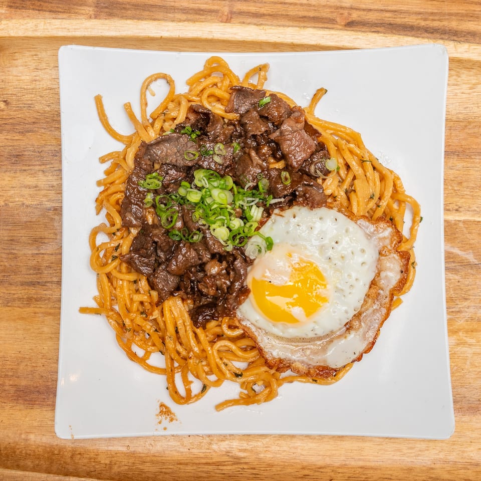 Orange noodles topped with sliced beef, green onions, and a sunny-side-up egg on a white plate atop a wooden table.