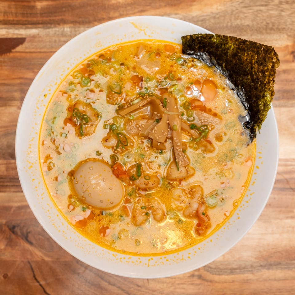 Overhead view of creamy spicy ramen topped with shrimp, egg, bamboo shoots, scallions, sesame seeds, and seaweed.