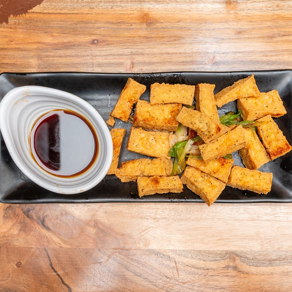 Crispy golden tofu pieces with sautéed vegetables and dipping sauce on a black rectangular platter.