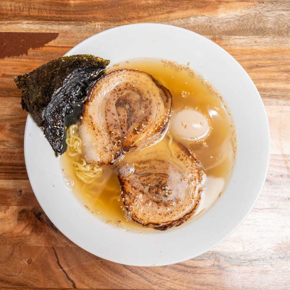 Overhead view of ramen in broth with noodles, sliced pork, a soft-boiled egg, seaweed, and sesame seeds on a wooden table.