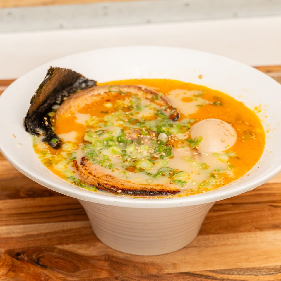 Bowl of creamy ramen topped with sliced pork, soft-boiled egg, green onions, sesame seeds, and seaweed in orange broth.